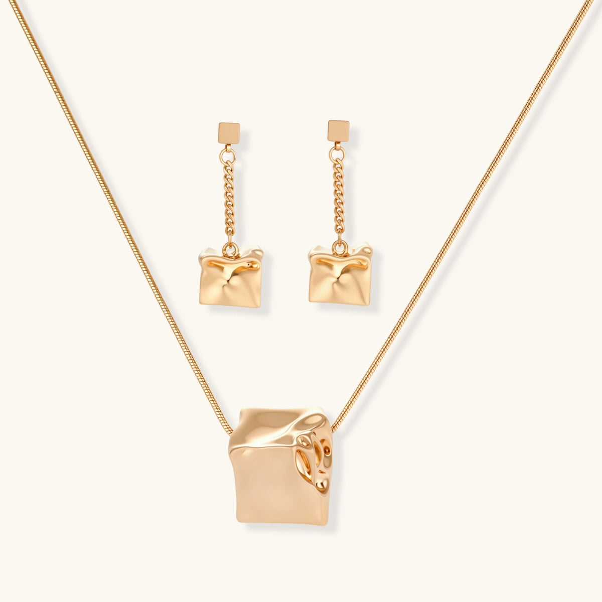 Modern geometric jewelry set featuring crumpled cube pendants in warm gold finish with chain details on beige background.